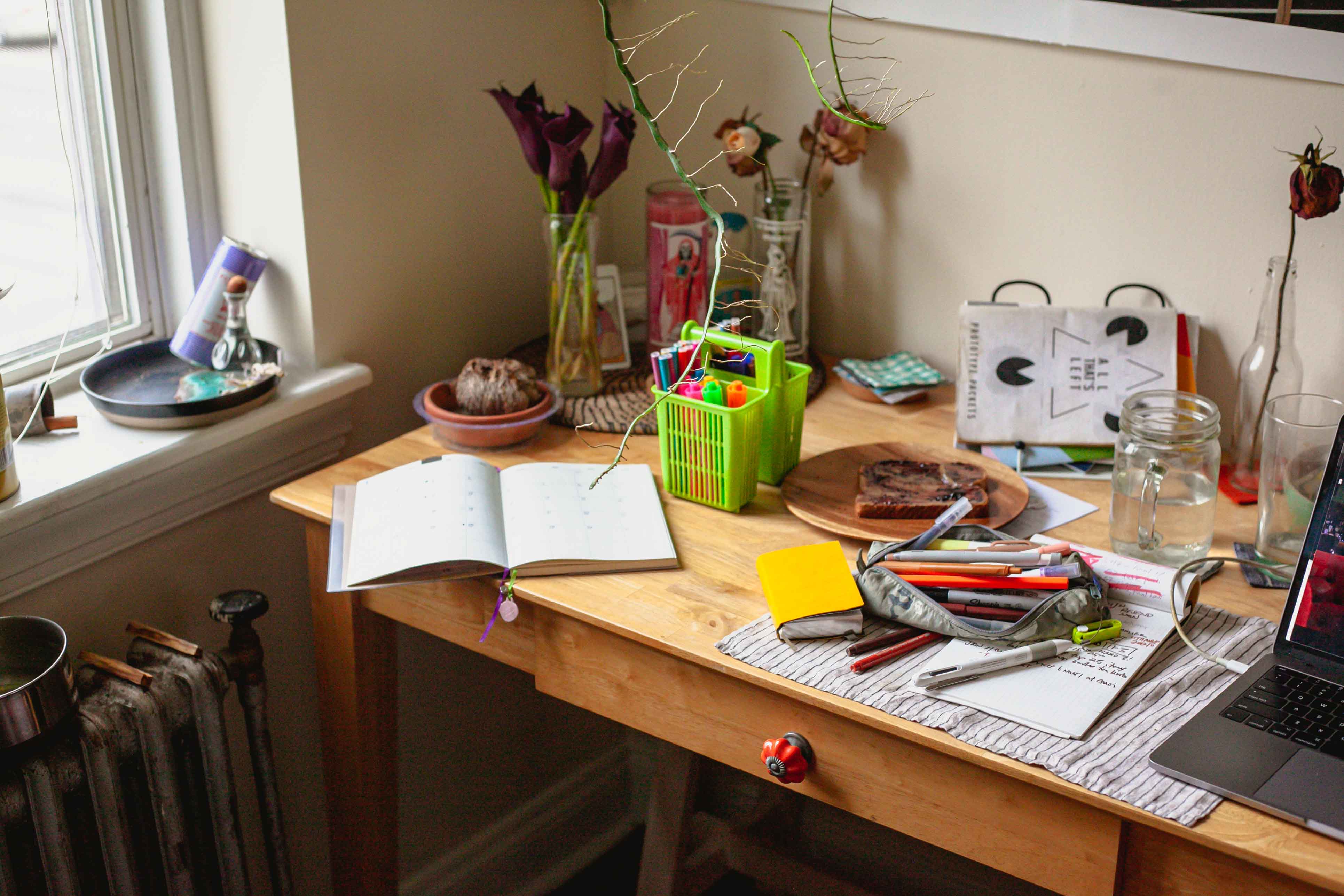 desk by a window, strewn with notebooks, zines, writing supplies.